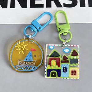 May include: Two keychains with colorful designs. One is a yellow circle with a boat and sun illustration, the other is a square with a house illustration. Both have metal clasps in blue and green.