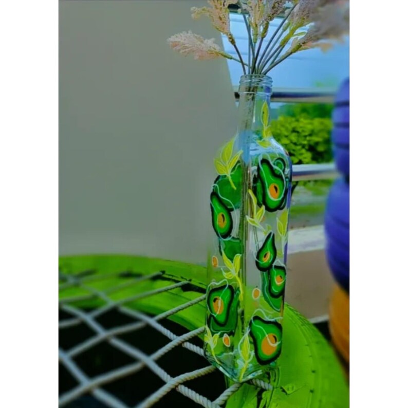 Puede incluir: Un jarr&oacute;n de botella de vidrio transparente decorado con dise&ntilde;os de aguacate verde y detalles de hojas amarillas. El jarr&oacute;n contiene un manojo de flores secas de color claro. El jarr&oacute;n est&aacute; sobre una superficie verde.