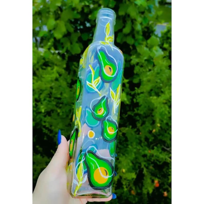 Puede incluir: Una botella de vidrio transparente decorada con dise&ntilde;os de aguacates pintados a mano. Los aguacates son verdes con centros amarillos y contornos negros. Acentos azules y amarillos y dise&ntilde;os de hojas completan la obra de arte. La botella se sostiene contra un fondo de follaje verde.