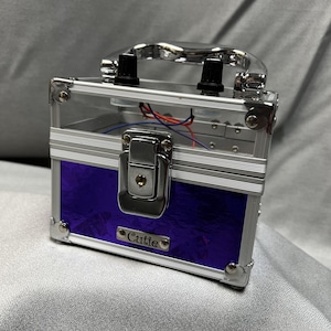 May include: A small, clear-sided case with a silver frame and a purple, textured front panel. The case has a silver handle, corner protectors, and a latch. The word "Cutie" is on a small silver plate on the front.