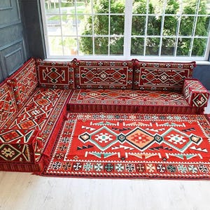 Puede incluir: Un conjunto de sofá de suelo en forma de L de tela roja con intrincados patrones geométricos en blanco, negro y verde azulado. El conjunto incluye varios cojines de respaldo, reposabrazos y una alfombra rectangular a juego con flecos.