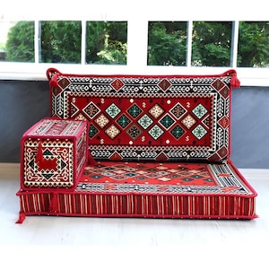 Puede incluir: Un conjunto de asientos de suelo que incluye un cojín rectangular, un respaldo y un reposabrazos. La tela es roja con patrones geométricos de diamantes en negro, blanco y verde y bordes a rayas, acabado con borlas rojas en las esquinas.