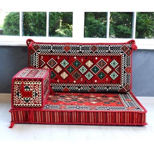 Puede incluir: Un conjunto de asientos de suelo que incluye un cojín rectangular, un respaldo alto y un cojín para el brazo. La tela es roja con patrones geométricos en negro, blanco y verde, con ribetes rojos y pequeñas borlas en las esquinas.