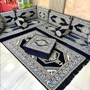 May include: A dark blue floor seating set with intricate geometric patterns in white, beige, and orange. The set includes cushions, a square ottoman, and a fringed rug, creating a cozy and stylish space.