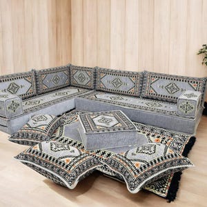 May include: A gray sectional sofa with matching patterned cushions and a square ottoman. The cushions and ottoman feature a geometric design with black, orange, and white accents. A coordinating rug with a similar pattern completes the set.