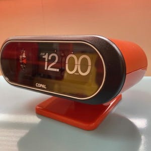 May include: A vintage orange and black Copal flip clock displaying 12:00 AM. The clock has a rectangular shape with a curved top and a square base. The word "Copal" is printed on the clock face.