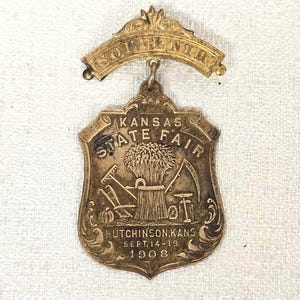 Puede incluir: Un pin de recuerdo dorado vintage de la Feria Estatal de Kansas, 1908. El pin presenta una forma de escudo con imágenes agrícolas y el texto "Kansas State Fair" y "Hutchinson, Kans. Sept. 14-19 1908". Una pancarta arriba dice "Souvenir".