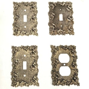 Puede incluir: Cuatro cubiertas de interruptores y enchufes de color latón antiguo. Cada cubierta es rectangular con un diseño floral y de hojas alrededor de los bordes. Dos cubiertas tienen aberturas para un solo interruptor, una tiene una abertura doble y otra tiene una abertura estándar para enchufe.