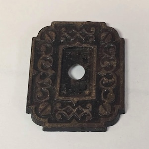 Op de afbeelding: Een donkerbruine metalen plaat met een ingewikkeld ontwerp rond de randen. De plaat heeft een rond gat in het midden.