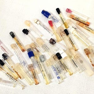 May include: A collection of small, clear perfume sample vials with various colored caps. Some vials have labels with brand names like CHANEL, Eternity, and Gucci. The vials are scattered on a white surface.