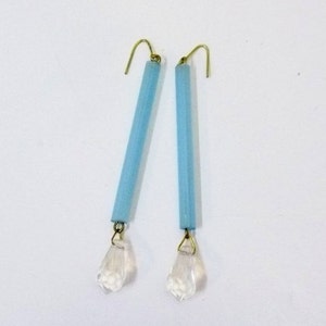 May include: A pair of dangle earrings featuring light blue cylindrical beads with gold-colored hooks. Each earring has a clear, faceted crystal teardrop charm suspended from the bottom. The earrings are set against a white background.
