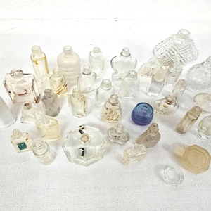 May include: A selection of small, clear glass bottles and vials in various shapes and sizes. Some have stoppers, others are open. One bottle is blue. The bottles are arranged on a white surface.