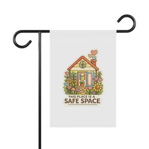 Peut inclure: Un drapeau de jardin blanc sur un support en métal noir. Le drapeau présente une illustration colorée d'une maison avec une porte arc-en-ciel, entourée de fleurs, et le texte "THIS PLACE IS A SAFE SPACE."