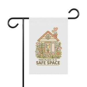 Peut inclure: Un drapeau de jardin blanc avec une illustration de maison et le texte "THIS PLACE IS A SAFE SPACE." La maison est entourée de fleurs et de cœurs. Le drapeau est fixé à un piquet de jardin en métal noir.