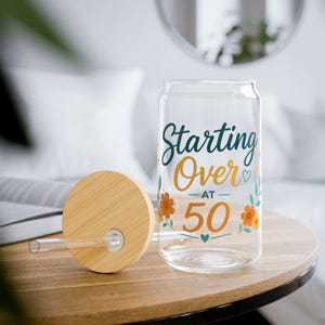Peut inclure: Un verre transparent avec un couvercle en bambou et une paille en verre. Le verre porte l'inscription Starting Over AT 50 en écriture bleu sarcelle et dorée, ornée d'illustrations florales orange, d'étoiles dorées et de cœurs bleu sarcelle.