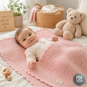 May include: A baby lies on a pink, scalloped-edge blanket. The baby is wearing a white onesie. A wooden sign with the words "you are so loved" is visible, along with a teddy bear and a wooden toy. A PDF pattern in English is also visible.