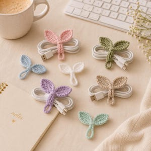 May include: A collection of handmade, crochet cable organizers in various pastel colors, shaped like leaves. Each organizer is wrapped around a white USB cable, offering a decorative and functional way to manage cords. The image includes a coffee cup, keyboard, and notebook.