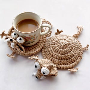 Puede incluir: Un juego de posavasos de ganchillo beige con un diseño de monstruo. Un posavasos sostiene una taza de café. El juego incluye dos posavasos adicionales. Los posavasos tienen ojos blancos grandes, pies pequeños y una boca detallada.