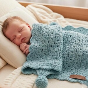 May include: A sleeping baby lies on a pillow, covered by a pale blue crocheted blanket. The blanket features a scalloped edge and a pom-pom detail. A small tag on the blanket reads "Handmade with Love".