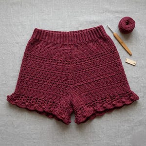 May include: Burgundy crochet shorts with a scalloped hem. The handmade shorts feature a textured stitch pattern and a comfortable waistband. A ball of yarn, a crochet hook, and a small wooden tag are next to the shorts.