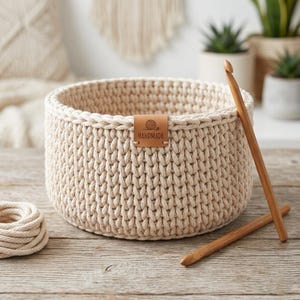 May include: A cream-coloured, handmade crochet basket with a leather tag reading "HANDMADE." Two wooden crochet hooks are propped against the basket. A ball of yarn sits to the left. The basket is on a wooden surface.
