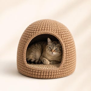 May include: A tan crochet cat bed shaped like a dome. A gray tabby cat is inside the bed, looking at the camera. The bed has a round opening and a soft, light-colored interior.