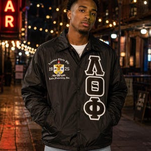 Puede incluir: Una chaqueta cortavientos negra con botones que presenta el escudo de Lambda Theta Phi en el pecho derecho con el texto 1925 y Latin Fraternity, Inc. El lado izquierdo muestra grandes letras griegas blancas A, O y Φ verticalmente.