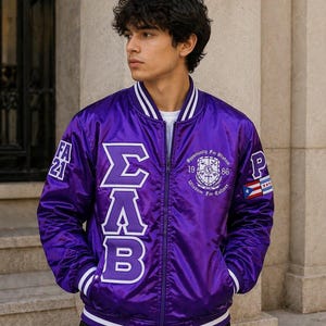 Puede incluir: Una chaqueta bomber de satén morado con ribetes de rayas blancas en el cuello, los puños y el dobladillo. El frente muestra las letras griegas Sigma, Lambda y Beta en blanco, junto a un escudo circular con el texto Opportunity for Wisdom 1986.