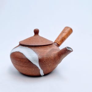 Peut inclure: Une théière en céramique brune avec une surface texturée et tachetée, dotée d'une poignée latérale. La théière présente un couvercle conique avec un petit bouton et un accent de glaçure blanche qui traverse le corps. Le bec est court et effilé.