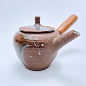Könnte beinhalten: Eine handgefertigte Keramik-Teekanne mit Holzgriff. Die Teekanne hat eine warme braune Farbe mit weißen und blauen Glasurakzenten. Der Deckel hat einen kleinen Knopf. Der Ausguss ist abgewinkelt und der Griff ist an der Seite befestigt.