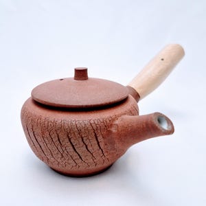 May include: A reddish-brown ceramic teapot with a wooden handle. The teapot has a lid and a spout. The surface of the teapot has a textured, cracked appearance. The handle is made of light-coloured wood.
