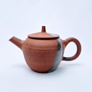Può includere: Una piccola teiera rotonda in ceramica marrone-rossastra con coperchio abbinato. La teiera ha un beccuccio curvo e un manico arrotondato. Una sezione verticale della teiera è di colore grigio scuro. Il coperchio ha una striscia bianca. La teiera è su sfondo bianco.
