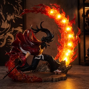 May include: A detailed figurine of a character in a dynamic pose, surrounded by vibrant, illuminated flames. The figure wears a red cape and black pants, with a sword at the ready. The flames are a bright orange and red, creating a dramatic effect.