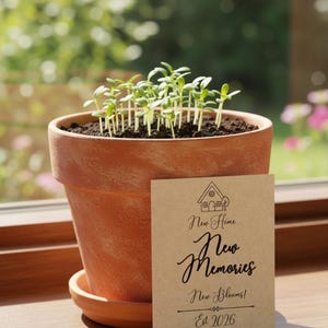 May include: A terracotta plant pot with young seedlings growing inside. A small card with the text "New Home, New Memories, New Blooms! Est 2026" is placed in front of the pot. The background shows a blurred view of a garden.
