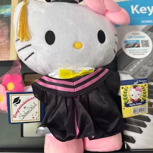 May include: A white Hello Kitty plush toy wearing a black graduation gown with pink trim, a matching mortarboard, and a yellow bow tie. The plushie has a pink bow and is holding a small graduation card.