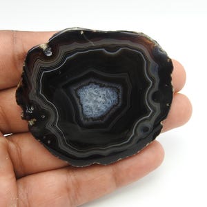 Può includere: Una fetta di agata lucidata con bande concentriche di nero profondo e grigio antracite, che circondano un ammasso centrale di quarzo cristallino bianco traslucido. La pietra presenta una superficie liscia e piatta con un bordo naturale.