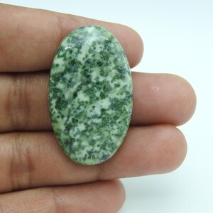 May include: An oval-shaped cabochon gemstone featuring a polished surface with a mottled pattern of deep forest green and creamy white speckles. The stone has a smooth, rounded dome and a flat base, displaying a natural, variegated mineral texture.