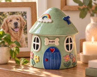 May include: A light green ceramic house-shaped container with a removable roof. The front features a blue door with a red heart, two arched windows, a bone-shaped plaque with the text BABY, a rainbow, a blue bow, and colourful flower accents.