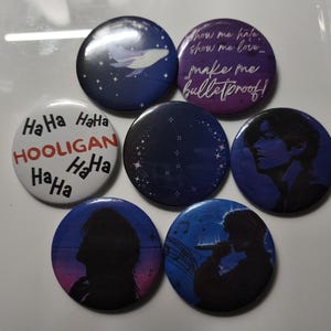 Könnte beinhalten: Eine Sammlung von sieben runden Ansteckbuttons mit verschiedenen Motiven. Ein Button ist weiß mit schwarzem Text HaHa und rotem Text HOOLIGAN. Andere zeigen dunkelblaue und violette Themen mit einem Wal, Sternbildern, einer menschlichen Silhouette und dem Text show me hale, show me love... make me bulletproof!
