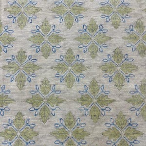 May include: A woven rug with a repeating pattern of green floral shapes and blue accents on a neutral background. The rug has a blue border along the bottom edge. The design is symmetrical and detailed.