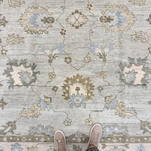 Könnte beinhalten: Ein hellblauer Teppich mit einem sich wiederholenden Blumenmuster in Beige-, Braun- und sanften Blautönen. Das Design umfasst symmetrische botanische Motive und komplexe, rankenartige Details, die über die gesamte Teppichfläche verteilt sind.