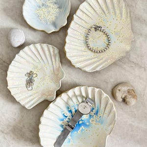May include: Four ceramic shell-shaped dishes with iridescent glazes in shades of blue, cream, and yellow. One dish holds a silver beaded bracelet, another a silver chain necklace, and a third a silver wristwatch with a mesh band.