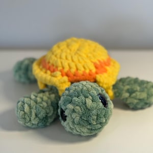 May include: A handmade crochet turtle plushie. The turtle has a yellow shell with orange stripes, green flipper-like appendages, and a green head with black safety eyes. The plushie is made of soft, textured yarn.