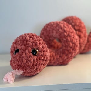 May include: A handmade, plush snake toy in a dusty rose color. The snake has large, shiny black eyes and a pink tongue. The body is coiled, and the soft texture is visible. This cuddly toy is perfect for children.