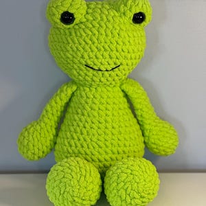 May include: A handmade, lime green crochet frog plush toy. The frog has large, black button eyes, a smiling mouth, and rounded limbs. The plush is made with a chunky yarn, giving it a textured appearance. The frog is sitting upright.