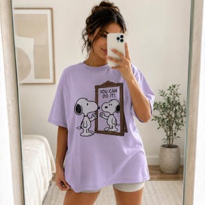 May include: A light purple short-sleeved t-shirt featuring a graphic of the cartoon character Snoopy looking into a mirror. The mirror frame is brown, and the text inside the mirror reads "YOU CAN DO IT!" in black capital letters.