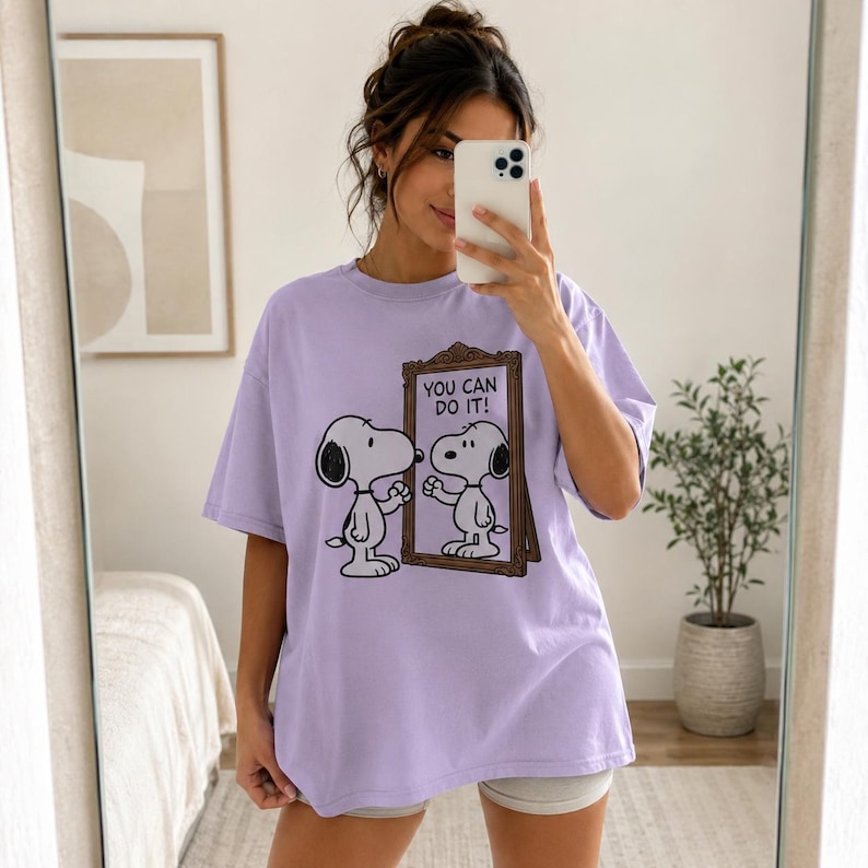 May include: A light purple short-sleeved t-shirt featuring a graphic of the cartoon character Snoopy looking into a mirror. The mirror frame is brown, and the text inside the mirror reads "YOU CAN DO IT!" in black capital letters.