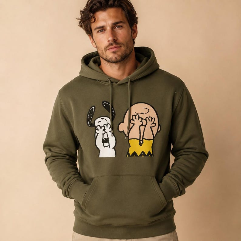 May include: An olive green hooded sweatshirt featuring a graphic print of Snoopy and Charlie Brown covering their eyes with their hands. The hoodie includes a front kangaroo pocket, long sleeves, and matching drawstring cords at the neckline.