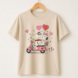 May include: A beige short-sleeved t-shirt featuring a graphic of two cartoon dogs riding a pink scooter. The design includes red and pink hearts, small stars, and a basket containing a letter, all rendered in a hand-drawn style on the front.