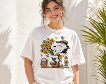 Könnte beinhalten: Weißes T-Shirt mit einem Cartoon-Snoopy-Design. Snoopy hält gelbe Blumen, trägt einen braunen Hut, Stiefel und einen gelben Schal. Ein Blumenkorb und weitere Blumen sind ebenfalls im Design enthalten.